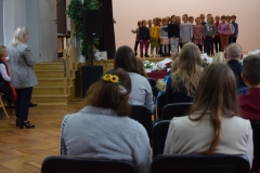 Na ciemnych krzesełkach w sali GCK siedzą widzowie oglądający występ dzieci. Przed nimi na scenie stoi grupa kilkunastu przedszkolaków w kolorowych strojach.     Z lewej strony widać dwie kobiety. Jedna z nich stoi. Ubrana jest w kraciastą marynarkę i ciemne spodnie. Druga z nich siedzi.