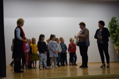 Na scenie stoi grupa dzieci przedszkolnych w kolorowych strojach. Z prawej strony stoją dwie kobiety w ciemnych spodniach i wzorzystych bluzkach. Jedna z nich trzyma mikrofon, druga dyplom i jasną reklamówkę. Po lewej stronie zwrócona twarzą do dzieci stoi trzecia kobieta ubrana w biało czarny strój i czerwony szalik.