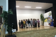 Na oświetlonej scenie stoi grupa kilkunastu przedszkolaków w kolorowych strojach. Po prawej stronie widać baner reklamujący gminę Wietrzychowice, z lewej urządzenie nagłaśniające, za nim sztuczny kwiat liściasty