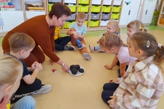 Dzieci siedzą w półkolu na podłodze, pani pokazuje im węgiel, w tle widać pudełko z zapałkami.