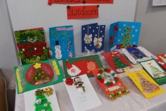 1. Na stolik nakryty białym obrusem wyłożone jest 14 kartek świątecznych. Widnieją na nich motywy choinek, bałwanków, mikołajów oraz różne elementy dekoracyjne. Na jednej z kartek widoczny napis czerwonymi, ozdobnymi literami: Wesołych świąt. Za tło służy biała tablica z czarnym napisem na czerwonych paskach kartonu Konkurs na kartkę świąteczną. Widok z perspektywy lewej strony.