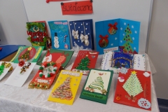 2. Na stolik nakryty białym obrusem wyłożone jest 14 kartek świątecznych. Widnieją na nich motywy choinek, bałwanków, mikołajów oraz różne elementy dekoracyjne. Na jednej z kartek widoczny napis czerwonymi, ozdobnymi literami: Wesołych świąt. Za tło służy biała tablica z czarnym napisem na czerwonych paskach kartonu Konkurs na kartkę świąteczną. Widok z perspektywy prawej strony.