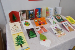 4. Na stolik nakryty białym obrusem wyłożone jest 17 kartek świątecznych. Widnieją na nich motywy choinek, bałwanków, mikołaj, renifer oraz różne elementy dekoracyjne. Na kilku kartkach  widoczny napis : Wesołych świąt.  Za tło służy biała tablica z czarnym napisem na czerwonych paskach kartonu Konkurs na kartkę świąteczną. Widok z perspektywy prawej strony.