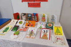 3. Na stolik nakryty białym obrusem wyłożone jest 17 kartek świątecznych. Widnieją na nich motywy choinek, bałwanków, mikołaj, renifer oraz różne elementy dekoracyjne. Na kilku kartkach  widoczny napis : Wesołych świąt. W tle biała tablica z fragmentem czarnego  napisu na czerwonym pasku kartonu Konkurs na kartkę świąteczną’. Widok z perspektywy lewej strony.