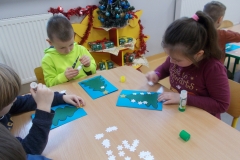 1. Przy stoliku w sali przedszkolnej siedzi czworo dzieci. Wyklejają one kartkę świąteczną dla rodziców: zielona choinka na niebieskim tle, na niej kompozycja           z białych śnieżynek, które leżą rozsypane na stole. Z prawej strony widać kolejny stolik i dziecko wykonujące to samo zadanie. W tle żółta szafka, na niej ubrana na niebiesko choinka, obok niej ,,prezenty’’ - pudełka opakowane w kolorowy papier       i przewiązane kokardką. Całość ozdobiona czerwonymi łańcuchami.