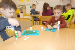 Przy stoliku przedszkolnym siedzi dwóch chłopców. Wklejają oni białe śnieżynki na zielone choinki. Znajdują się one na niebieskich kartkach papieru. Za nimi widoczny stolik z czterema chłopcami, którzy wykonują taką samą pracę. W tle szafki przedszkolne.