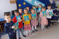 3. Grupa odświętnie ubranych przedszkolaków stoi obok choinki. Dzieci trzymają w rękach wykonane przez siebie kartki świąteczne dla rodziców. Mają one kolor niebieski, na nich są zielone choinki ozdobione białymi śnieżynkami i żółtą gwiazdką na czubku. Na dole, na żółtym tle napis ,, Wesołych świąt’’. Dzieci stoją na tle granatowej dekoracji, na której, na niebieskiej wstędze znajduje się napis,, Cicha noc’’ białymi, drukowanymi literami. Zdobią ją też żółto - czerwone gwiazdki.