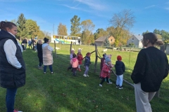 W zagrodzie z drewnianych pali i siatki po zielonej trawie chodzą kozy małe i duże, białe, czarne i brązowe. Przy siatce stoją dzieci. Nieco dalej panie i panowie. W oddali na drodze stoi biały autobus szkolny z niebieskim, zielonym i żółtym pasem. Za drogą widać domy.