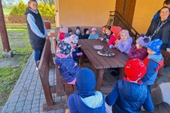 Na szarej kostce stoi brązowy, prostokątny stół. Na nim  talerz z ciastkami i szklana misa z cukierkami. Wokół stołu ustawione brązowe ławki, na których siedzą dzieci. Po lewej stronie stoi pani w niebieskim dresie i czarnej kamizelce. Po prawej pani w czarnej kurtce, a za jej plecami pan w granatowej kurtce. W tle żółty budynek z brązowymi drzwiami.