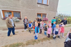 Dzieci stoją na drodze wysypanej kamieniem. Przed nimi pan w brązowej bluzie. Obok dzieci 4 panie. Jedna trzyma w rękach koszyk z kwiatami. W tle szary dom. Na 2 parapetach skrzynki z białymi, różowymi i fioletowymi kwiatami.