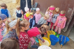 Dzieci w kolorowych ubraniach stoją w grupie i uważnie się przyglądają. 2 dziewczynki pochylone mieszają mleko w żółtym wiaderku. Obok stoją niebieski i żółte wiadra ze smoczkami. Za dziećmi brązowa ściana.