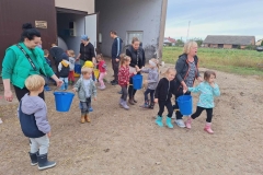 3 panie niosą 3 niebieski wiadra. Przy każdej z nich po 2 dzieci. Za nimi pan i pani z żółtym wiadrem, które pomaga nieść dziewczynka w różowych spodniach. Tyłem odwrócona pani w czarnej bluzie. Obok niej 4 dzieci.