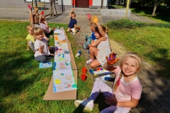 Dzieci siedzą w dwóch rzędach, pomiędzy nimi białe kartki, stemplują rączkami szal dla pani jesieni.