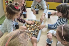 Dzieci siedzą przy stoliku, segregują do pojemników różne elementy.