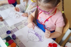 Na zdjęciu widać dwie dziewczynki, siedzą przy stoliku, malują patyczkami kosmetycznymi statek kosmiczny. Jedna z nich ma okulary.