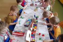 Dzieci siedzą przy złączonych stolikach, po obu stronach, dzieci wykonują prace plastyczną techniką puentylizm. Na stole leżą farby plakatowe.