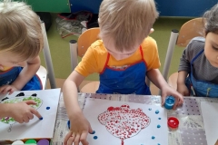 Chłopiec w niebieskim fartuszku maluję na kartce niebo wokół statku kosmicznego opuszkami palców.