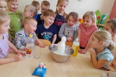 Dzieci stoją wokół stołu, pani wsypuje sodę do butelki umieszczonej w misce.