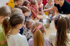 Dzieci stoją wokół stołu, z butelki umieszczonej w misce wydobywa się białą piana.na stole leży ocet, soda i płyń do naczyń.