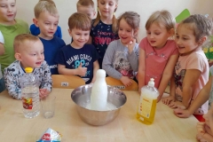 Dzieci stoją wokół stołu, z butelki umieszczonej w misce wydobywa się białą piana.