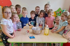 Grupa dzieci stoi wokół stołu. Na stole leży miska, płyń do naczyń, ocet i soda oczyszczona.