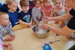 Dzieci stoją wokół stołu  pani wlewaliśmy do butelki umieszczonej w misce ocet.