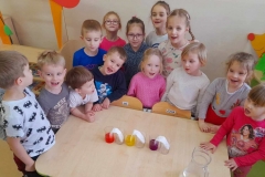 Dzieci zgromadzone wokół stolika na którym stoi 6 szklanek. W 3 zabarwiona wodą, 3, do nich włożone papierowe ręczniki i 3 puste.