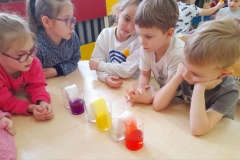 Na stole leży 6 szklanek, 3 puste, w 3 zabarwiona wodą bibułą. Pani wyciąga z ostatniej szklanki fioletową bibułę.