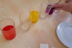 Na stole leży 6 szklanek, 3 puste, w 3 zabarwiona wodą bibułą. Pani wyciąga z ostatniej szklanki fioletową bibułę.