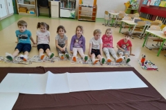 Dzieci siedzą na podłodze, na stopach mają nałożona folie babelkowa. Stopy pomalowane są kolorowa farba. Przed dziećmi bialy tor z kartek ułożony na brązowym materiale.