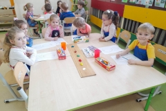 Przy stoliku siedzą dzieci, na stole widać farby oraz kolorowy gwasz na deseczce. Przed dziećmi kartki z wiosennymi kwiatami, które będą malować.