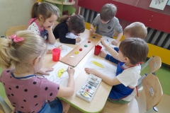 Przy stoliku siedzi grupa dzieci, maluję wiosenne kwiaty kolorowym gwaszem, który widać na deseczce.