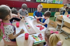 Przy stoliku siedzą dzieci, malują gwaszem, na kartkach widać kolorowe motyle.