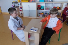 Na środku sali przedszkolnej ustawione zostało małe, jasne biurko. Leży na nim notes i długopis. Za biurkiem siedzi chłopiec ubrany w biały fartuch, na szyi ma zawieszone lekarskie słuchawki. Przed biurkiem siedzi chłopiec pacjent, ubrany w ciemne spodnie, białą koszulkę i rozpiętą pomarańczową bluzę. W tle widoczne meble przedszkolne, na nich pudełka z klockami.