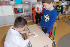 Za biurkiem siedzi chłopiec przebrany za lekarza i wypisuje receptę. Przed biurkiem stoi chłopiec w niebieskiej bluzie z nadrukowaną piłką. Za nimi w tyle widoczne są dzieci - pacjenci, stojący i siedzący na krzesełkach. W tle meble, na nich zabawki.