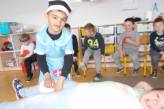 Na pierwszym planie widać postać leżącej, uśmiechniętej dziewczynki w jasnym ubraniu. Za nią stoi chłopiec w niebieskim  stroju pielęgniarki i biało - czarnej opasce na głowie. Bandażuje dziewczynce nogę. W tyle, na krzesełkach siedzą dzieci. Za nimi meble przedszkolne.