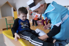 Na pierwszym planie widać postać leżącej, uśmiechniętej dziewczynki w jasnym ubraniu. Za nią stoi chłopiec w niebieskim  stroju pielęgniarki i biało - czarnej opasce na głowie. Bandażuje dziewczynce nogę. W tyle, na krzesełkach siedzą dzieci. Za nimi meble przedszkolne.