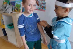 Chłopiec przebrany w niebieski strój pielęgniarki robi zastrzyk pacjentowi - chłopcu ubranemu w niebieską bluzę i zielone spodnie. Z boku widoczna szafka z książkami.