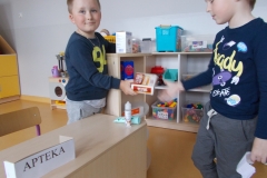 Na środku sali przedszkolnej stoi małe, jasne biurko. Z boku umieszczony jest napis Apteka, drukowanymi czarnymi literami na białym tle. Chłopiec ubrany w ciemną bluzę i jasne spodnie odgrywa rolę aptekarza - podaje pudełko z lekarstwami chłopcu w ciemnym ubraniu z nadrukiem. Trzyma on w ręku kartkę - receptę.