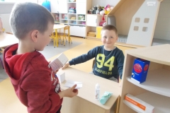 Za biurkiem siedzi chłopiec w ciemnym ubraniu z żółtym napisem: 94 bluefields drukowanymi literami. Na boku biurka i stojącej obok szafce leżą opakowania po lekarstwach. Przed biurkiem stoi chłopiec w czerwonej bluzie, trzyma w ręku opakowanie lekarstw i receptę.