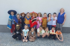 Grupa dzieci w kolorowych strojach stoi na tle białego budynku. Przed nimi na kostce klęczy lub kuca siedmioro dzieci. Obok stoi pani w szarych spodniach i niebieskiej bluzce.