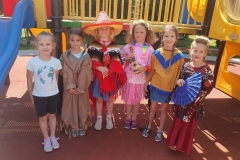 Dziewczynki przebrane za Indianki, Meksykankę, Hawajkę, Japonkę stoją przy niebieskiej zjeżdżalni. Japonka trzyma w ręku wachlarz, Hawajka ukulele, Meksykanka ma na głowie sombrero.