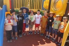 Chłopcy przebrani za dzieci z różnych stron świata stoją  w półkolu na placu zabaw. 2 trzyma w rękach bagietki, 2 ma na głowach kapelusze, 1 ciupagę, 1 maskę na twarzy.