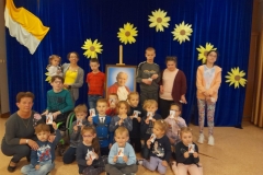 Grupa dzieci siedzi na podłodze, za nimi stoją dzieci z ośrodka, pośrodku dzieci portret papieża.