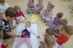 Dziewczynka leży na białym kartonie, pani rysuje jej kontur, wokoło siedzą dzieci.