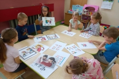 Dzieci siedzą przy stolikach, na stołach rozłożone rysunki: symbole agresji. Dziewczynka trzyma w rękach rysunek  opowiada o nim.