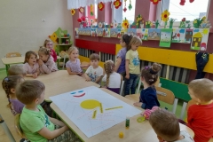 Dzieci siedzą przy stolikach, na stoliku biały karton  żółte koło, promyki słońca. Dwie dziewczynki stoją plecami do siebie, sprawdzają czy są równe.