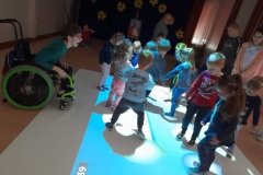 Grupa dzieci bawi się na magicznym dywanie, z boku widać chłopca na wózku inwalidzkim.