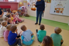 Dzieci siedzą w półkolu na podłodze. Pani dietetyk omawia im piramidę zdrowia, w ręku trzyma koszyk z mandarynkami.