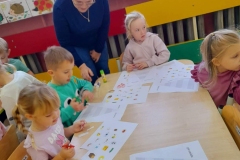 Dzieci siedzą przy stoliku, wyklejaja piramide, pani podpowiada jak to zrobić. 